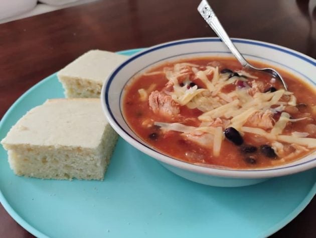 A bowl of three-bean chili topped with shredded cheese, served on a blue plate with two thick slices of cornbread.