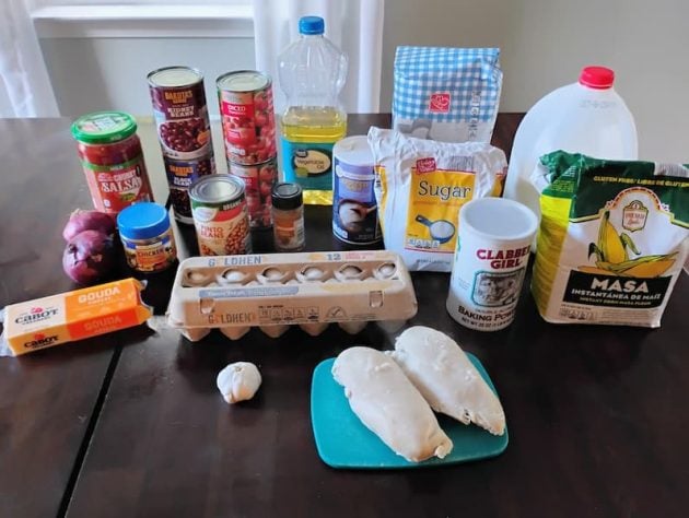 A variety of ingredients for a chili and cornbread recipe are laid out on a dark table,