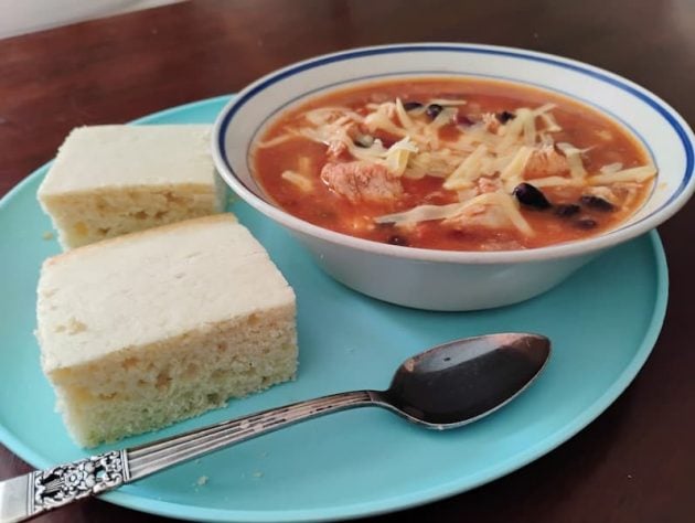 A bowl of three-bean chili chowder topped with shredded cheese, served on a blue plate with two thick slices of cornbread.