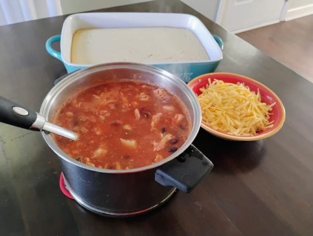 A pot of three-bean chili sits on a dark table, with a bowl of shredded cheese and a rectangular baking dish filled with freshly baked cornbread.