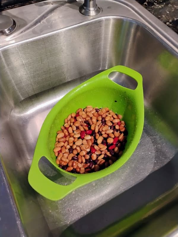 A green colander is filled with beans in a stainless steel sink.