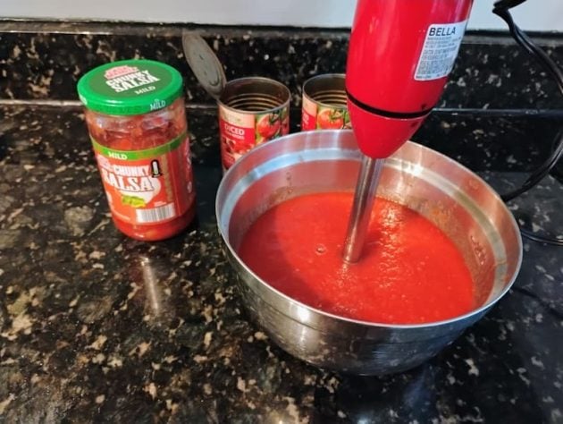 A red immersion blender is mixing a bowl of blended tomatoes on a black granite countertop for chili.