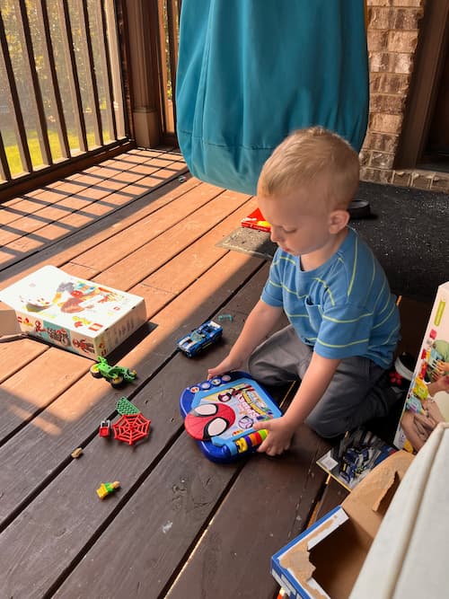 Young boy sits on a sunny wooden porch floor playing independently with a drawing tablet and scattered toys while parents work or homeschool nearby.
