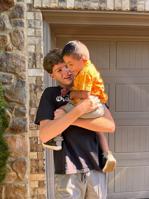 Teenage boy stands outside a brick home, smiling warmly as he holds a younger child dressed in an orange shirt who is laughing and snuggling close.