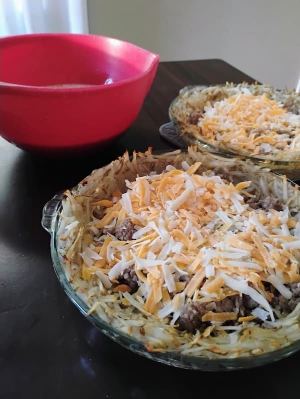 Two glass pie dishes filled with shredded potato crust, cooked sausage, and a generous layer of shredded cheddar jack cheese, sitting next to a red mixing bowl of beaten eggs, ready to be assembled into meat quiche with potato crust.