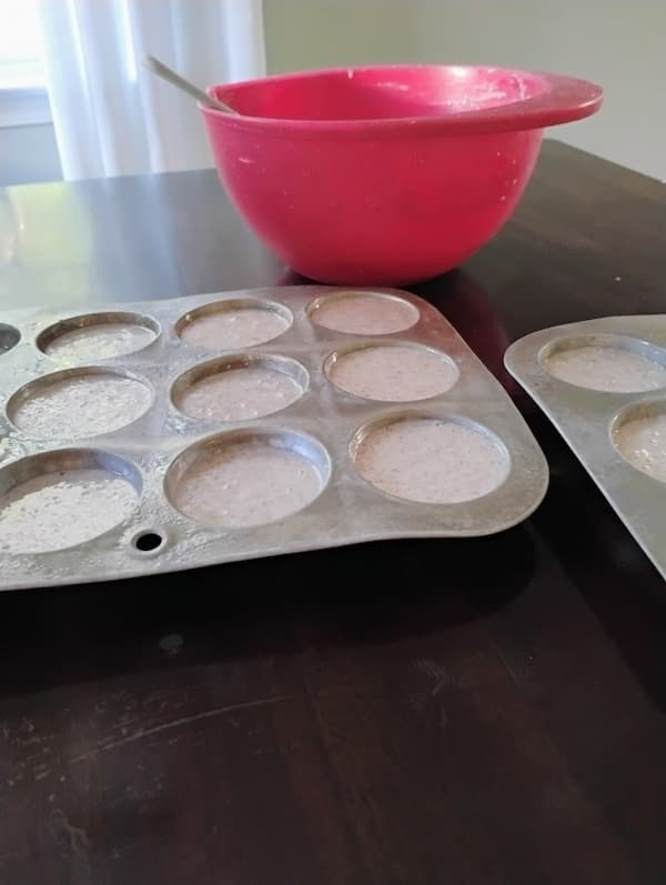 Muffin tins filled with flax muffin batter sit on a dark table next to a red mixing bowl with a spoon, ready to be baked.