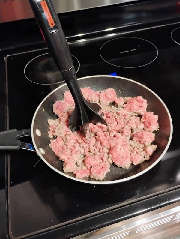 Ground pork sausage cooking in a skillet on a stovetop, being broken up with a meat chopper utensil as it browns.