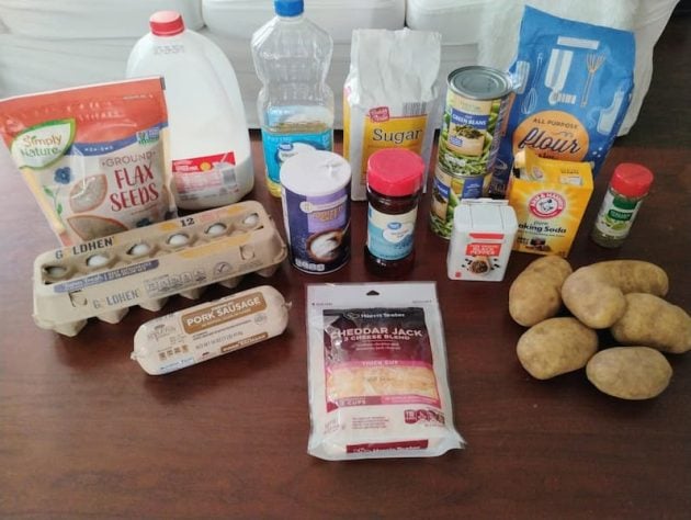 Ingredients laid out on a table for making meat quiche with potato crust and flax muffins, including eggs, shredded cheddar jack cheese, ground pork sausage, russet potatoes, milk, oil, flour, sugar, baking soda, baking powder, canned vegetables, spices, ground flax seeds, and jam.