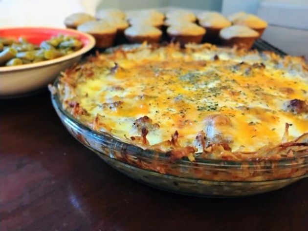 Golden-brown meat quiche with a crispy potato crust in a glass pie dish, topped with melted cheese and herbs. A bowl of green beans and a cooling rack of flax muffins sit in the background.