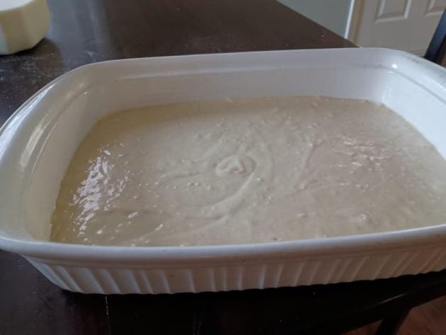 A white baking pan is filled with cornbread batter sitting on a kitchen table.
