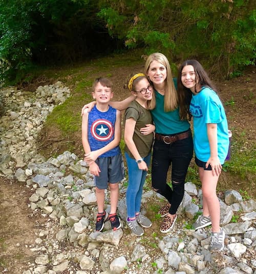 Woman standing on a rocky trail smiles and hugs three children—two girls and a boy—while surrounded by trees and greenery