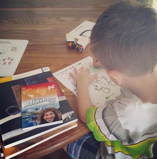 Young boy sits at a wooden table coloring an educational worksheet, surrounded by crayons and homeschool books showing a hands-on moment of learning, relevant to how to balance working from home and homeschooling.