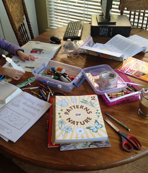 Round wooden table covered with homeschooling supplies including open books, coloring pages, scissors, markers, and a “Patterns of Nature” workbook, alongside a desktop computer and coffee mug. A child’s hands are visible, highlighting the multitasking reality of working from home and homeschooling.